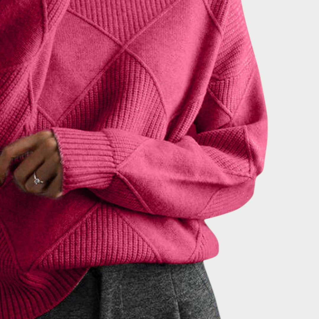 Zane & Inese Gaida pink knit sweater with a high collar and geometric diamond pattern, worn with grey trousers.