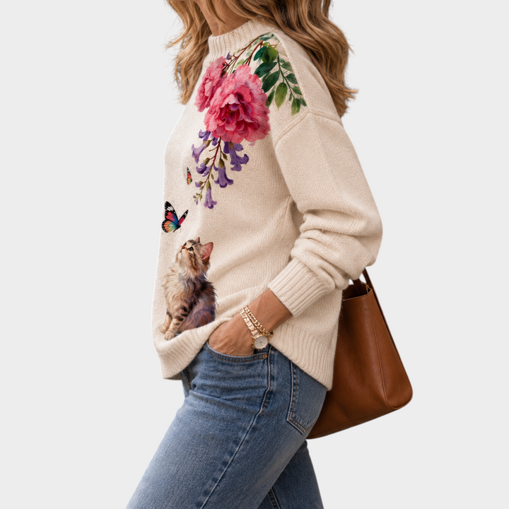 Zane & Inese cream knit sweater with high neck, floral embroidery, and a kitten graphic, worn with blue jeans.